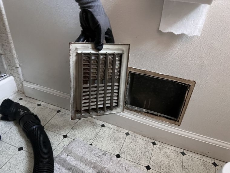 Technician's gloved hand holding a removed vent cover during air duct cleaning
